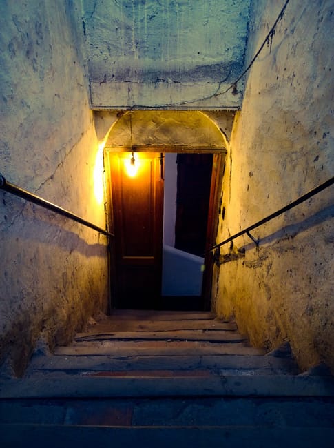 A narrow, steep staircase with aged, worn wooden steps and concrete walls leading down to a door at the bottom. The staircase is illuminated by a single exposed light bulb attached to the wall, castin