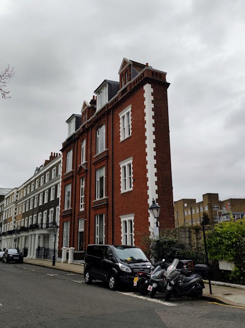 A multi-storey red brick building on a narrow street in South Kensington with classic white window frames and decorative white vertical stripes along the corner. In front, there is a black van parked on the street, with three black motor scooters and a helmet placed on the pavement nearby. The street is lined with other residential buildings, some painted white, and features a sidewalk with a black lamppost. Overcast grey clouds cover the sky, creating diffused natural light. This setting illustrates typical urban surroundings suitable for house removals or furniture transport services in the South Kensington SW7 area, with the building's architecture and street scene conveying a residential property ready for relocation or removal operations managed by companies like manandavansouthkensington.co.uk.