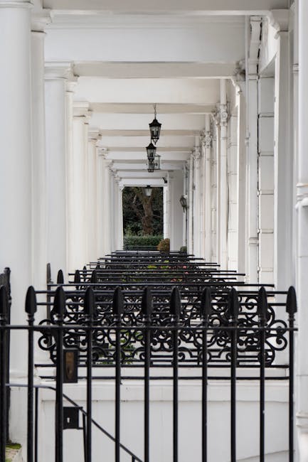 A covered walkway with white-painted columns and a ceiling, illuminated by hanging lantern-style light fixtures. Black wrought-iron railings with decorative scrollwork line the front of the porch, which encloses a small garden area visible beyond the railing. The pathway leads towards a tree with green foliage in the distance. This setting suggests a residential property in South Kensington, suitable for house removals or home relocation services, with the focus on the architectural details and entrance area that may be involved in moving furniture or packing materials during a house move.